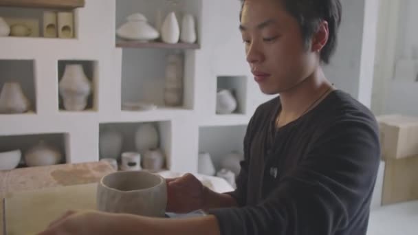 スタジオで働いている間に陶器の車輪に粘土花瓶をかけるためにスポンジと水を使用して若いアジアのセラミストの中型ショット — ストック動画