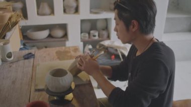 Çömlekçilik atölyesinde el yapımı kil vazoyu dekore ederken boya fırçası arayan genç Asyalı bir seramistin orta açı görüntüsü.