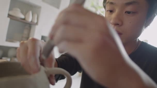 陶芸スタジオで粘土マグカップにハンドルを添付しながらポッターリブを使用した若いアジアのセラミストのラックフォーカスショット — ストック動画