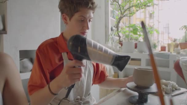 Junge Kurzhaarige Frau Schürze Trocknet Keramikbecher Auf Töpferscheibe Mit Haartrockner — Stockvideo
