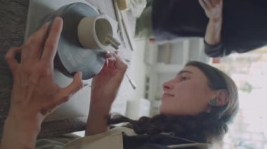Genç bir kadının çömlek çarkında çamurdan kupa yaparken ana zanaat sınıfında seramik yaparken dikey görüntüsü.