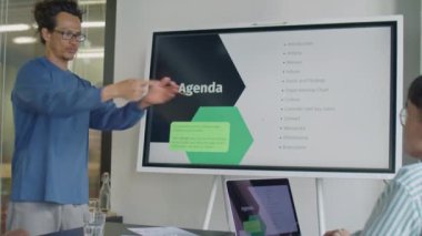 Asyalı adam dijital ekranda iş sunumu yapıyor ve ofis toplantısı sırasında çok ırklı meslektaşlarıyla gündemi tartışıyor