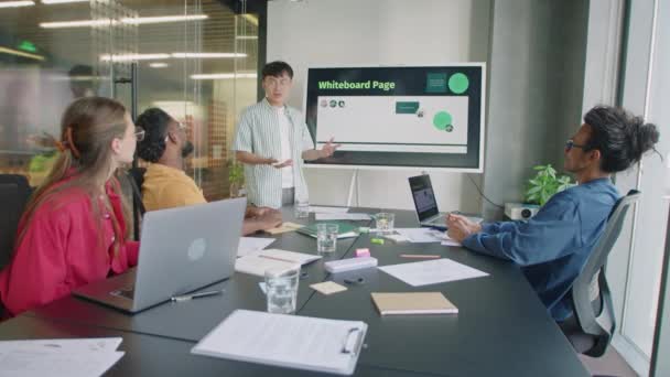 デジタルスクリーンで立ち上がり オフィスでのミーティング中にビジネスプレゼンテーションをしながらチームとコミュニケーションをとる若いアジア人男性 — ストック動画