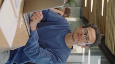 Asyalı bir adamın gündelik kıyafet ve gözlüklü portresi ofiste dizüstü bilgisayarla oturmuş gülümseyerek poz veriyor.