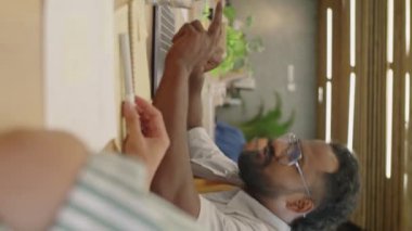 Genç Hintli adamın dizüstü bilgisayarı işaret edip iş projesini ofisteki meslektaşıyla tartışırken dikey görüntüsü.