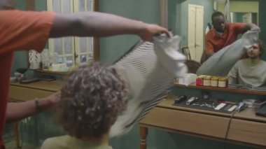 Genç kıvırcık saçlı adam aynanın önünde berber koltuğunda otururken siyah berber ona kuaförlük pelerini takıyor.