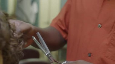 Berber dükkanında çalışırken erkek müşterinin kıvırcık saçlarını seyrelten makaslarla kesen genç siyah berberi yakından çek.