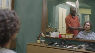 Berber dükkanında oturan genç adam aynaya bakıyor ve Afrikalı Amerikalı berberle saç kesimini tartışıyor.