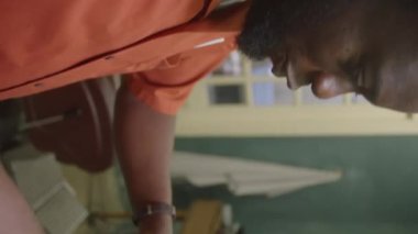 Dikey çekimde genç siyah berber erkek müşterinin kıvırcık saçlarını makasla kesiyor ve berber dükkanında çalışırken onunla konuşuyor.