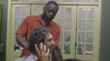 Kuaför pelerinli genç adam berber dükkanında oturup profesyonel siyah berbere nasıl bir saç kesimi istediğini anlatıyor.