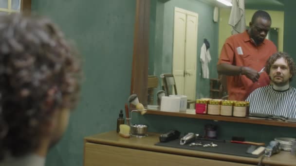 Młody Kręcone Włosy Mężczyzna Pelerynie Fryzjerskiej Siedzi Przed Lustrem Podczas — Wideo stockowe