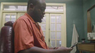 Afrika kökenli Amerikalı erkek berberin retro berber dükkanında otururken ve akıllı telefondan internette sörf yaparken çekilmiş bir fotoğrafı.