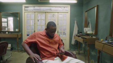 Orta boy Afro-Amerikan erkek berber koltukta otururken ve cep telefonuyla sosyal medyada kayarken vintage berber dükkanında mola verirken.