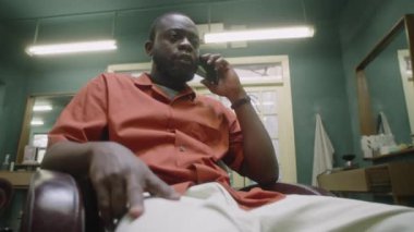 Siyah erkek berberin eski moda berber dükkanında oturduğu ve iş günü cep telefonuyla konuştuğu düşük açılı bir çekim.