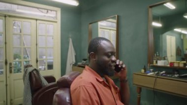 Eski moda berber dükkanında oturan ve cep telefonuyla konuşan Afrikalı Amerikalı erkek berberin orta boy fotoğrafı.