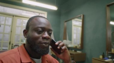 Kuaförde oturan ve cep telefonuyla konuşan sakallı Afrikalı Amerikalı bir erkek berber.
