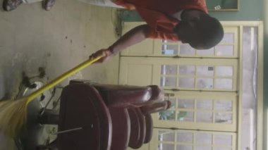 İş gününden sonra kuaför salonunu temizlerken, yerleri süpüren Afrikalı Amerikalı berberin dikey görüntüsü.