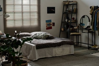 Gri battaniyeli rahat çift kişilik yatak ve pencerenin yanında duran yastıklar masa ve raflar arasında Venedik panjurlarıyla kaplı.