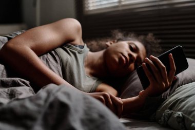 Yatakta uzanmış yeni siber zorbalık mesajlarına bakarken elinde akıllı telefon tutan stresli Afrikalı Amerikalı bir gencin eline odaklan.