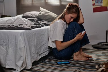 Hüsrana uğramış, gündelik giysili genç bir kız yerde, akıllı telefonun yanında oturmuş elinde haplarla şişeye bakıyor.