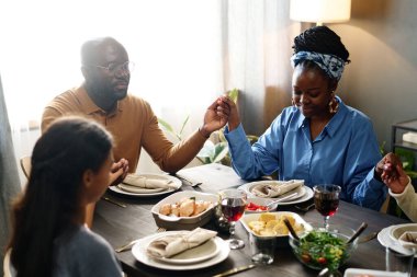 Şükran Günü yemeği için el ele tutuşan dört kişilik modern Afro-Amerikan ailesi yemek ve içeceklerle servis yaparken...