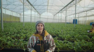 Geniş bir serada çilek sıraları arasında kameraya poz veren neşeli genç kadın.