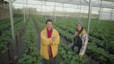 Geniş bir sera çiftliğinde kameraya poz veren ve gülümseyen iki genç bayanın yüksek açılı çekimi.