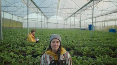 Genç kadın çiftlik işçisi kameraya bakıyor ve büyük serada dururken gülümsüyor. Meslektaşı arka planda böğürtlen topluyor.