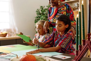 Kwanzaa gününde küçük kız kardeşi ve annesinin yanında otururken milli kostümlü şirin Afro-Amerikan kız.