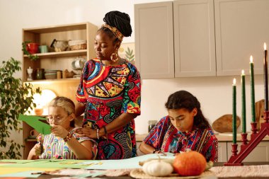 Young woman in national attire standing by her daughter cutting green paper while making greeting card for kwanzaa celebration at home clipart