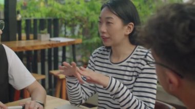 Genç Asyalı bayan takım lideri açık terasta yapılan toplantıda çok ırklı meslektaşlarına iş fikirlerini anlatıyor.