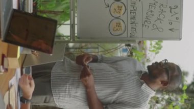 Siyah iş adamının beyaz tahtada dikey çekimi ve açık terasta yapılan toplantıda çok ırklı takımlara sunum yapması.