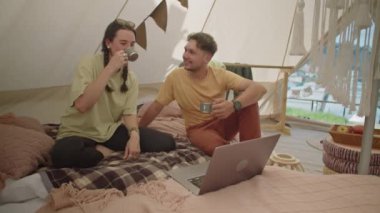 Genç çift çadırda birlikte oturuyor, çay içiyor, sohbet ediyor ve tatil sırasında internette sörf yapıyor.