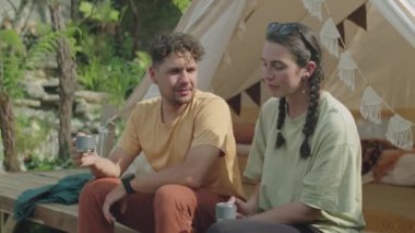 Genç çiftin çadırın dışında oturup çay içerken ve yaz tatilini birlikte geçirirken çevreyi tartışırken orta boy bir fotoğrafı.