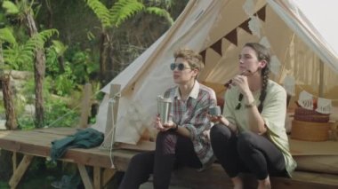 İki kadın turistin çadırın dışında oturup, çay içerken, doğaya bakarken ve yaz tatilinde sohbet ederken orta uzunlukta görüntüsü.