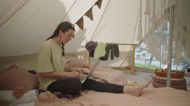 居心地の良いグランピングテントに床に座り 自然の中で休暇中にリモートで作業しながらラップトップにタイピングする若い女性観光客 — ストック動画