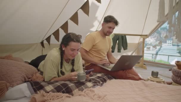 年轻夫妇一起躺在帐篷里 喝着茶 用智能手机 男人们用笔记本电脑在网上冲浪 — 图库视频影像
