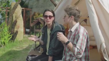 Glampsite 'de oturan, çay içen ve doğada tatilde sohbet eden iki genç kız arkadaşın orta boy fotoğrafı.