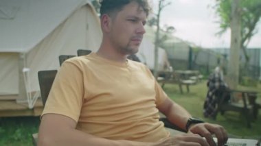 Erkek turistin Glampsite 'deki çadırın önünde otururken, dizüstü bilgisayarda yazı yazarken ve tatil sırasında doğaya bakarken görüntüsünü kaldır.