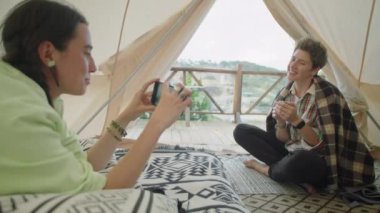 Genç bir kadın çadırda uzanıyor ve elinde çay fincanı tutarken akıllı telefon kullanıyor ve gülümsüyor.