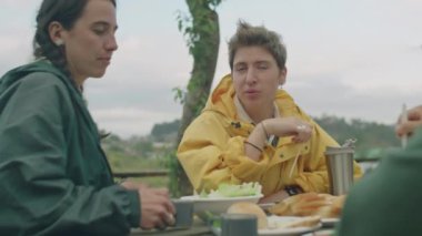 İki kız dışarıda masada oturuyor, yemek yiyor ve kamp alanında piknik yaparken arkadaşlarıyla sohbet ediyorlar.