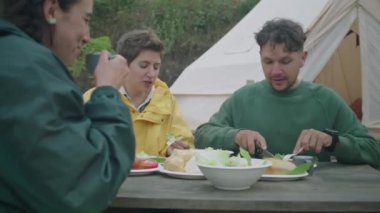 Orta halli turist topluluğu çadırın dışında yemek masasında oturuyor, yemek yiyor ve akşam yemeğinde konuşuyor.