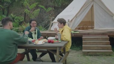 Orta ölçekli neşeli turistler çadırın dışında yemek masasında oturur, akşam yemeğinin tadını çıkarır ve hafta sonları doğada sohbet ederler.