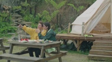İki kız arkadaşın açık havada çadırın önünde otururken ve doğada akşam yemeği yerken akıllı telefonla selfie çekerken.