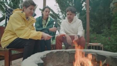 Neşeli turistlerle dolu orta ölçekli bir çekim. Kamp ateşinin yanında oturuyorlar, konuşuyorlar ve alevler üzerinde bbq sosisi pişiriyorlar.