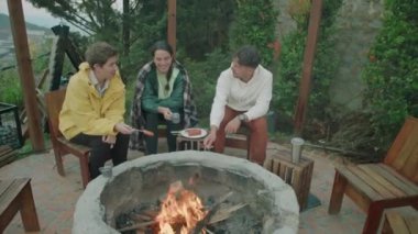 Genç turistlerin dağlık bölgede kamp ateşinin yanında otururken yüksek açılı çekimleri, sohbet ve alevler üzerinde bbq sosisleri pişirirken.