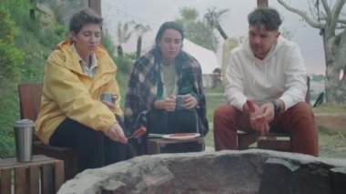Kamp ateşi üzerinde sosis pişiren ve doğada sohbet eden genç turistlerle dolu orta ölçekli bir çekim.
