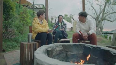 Glampsite 'de kamp ateşi başında oturmuş, çubuklarda ızgara sosis yapıp, dışarıda sohbet eden genç turistlerden oluşan orta ölçekli bir çekim.