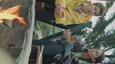 İki genç kadının kamp ateşi üzerinde sosis pişirirken ve doğada dinlenirken çay içerken sohbet ederken dikey çekimleri.