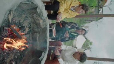 Kamp ateşinde dinlenen, ızgara sosis pişiren ve çay içen genç turistlerin dikey görüntüsü. Hafta sonunu doğada geçirirken bir şeyler hakkında konuşmak.
